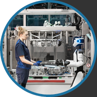 A factory worker in uniform interacts with a humanoid robot in a high-tech manufacturing setting. The scene conveys a collaborative and futuristic atmosphere.