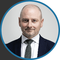 A man in a suit and tie poses professionally against a neutral gray background.