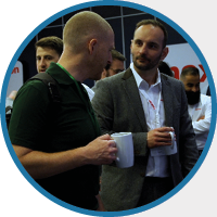 Two men holding mugs engage in conversation at a networking event. They're in business attire, surrounded by others in a lively, professional setting.