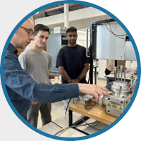 A group of four people stands in a lab setting, focused on a man demonstrating equipment on a wooden table. The mood is attentive and collaborative.