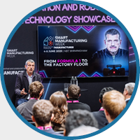 A speaker addresses an audience at the Smart Manufacturing and Robotics Technology Showcase. The presentation screen features the topic 'From Formula 1 to the Factory Floor.'