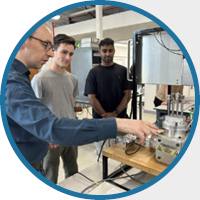 A man in a blue shirt demonstrates a lab machine to two attentive students. The room is filled with technical equipment, suggesting a scientific setting.